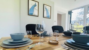 Une table à manger est dressée avec de l’assiette bleue, des bols et des verres à vin, et deux tableaux représentant des paons, encadrés, sont accrochés au mur.