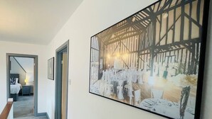 Un couloir orné d’un tableau représentant l’intérieur d’une grange, avec un lit et une table de chevet.