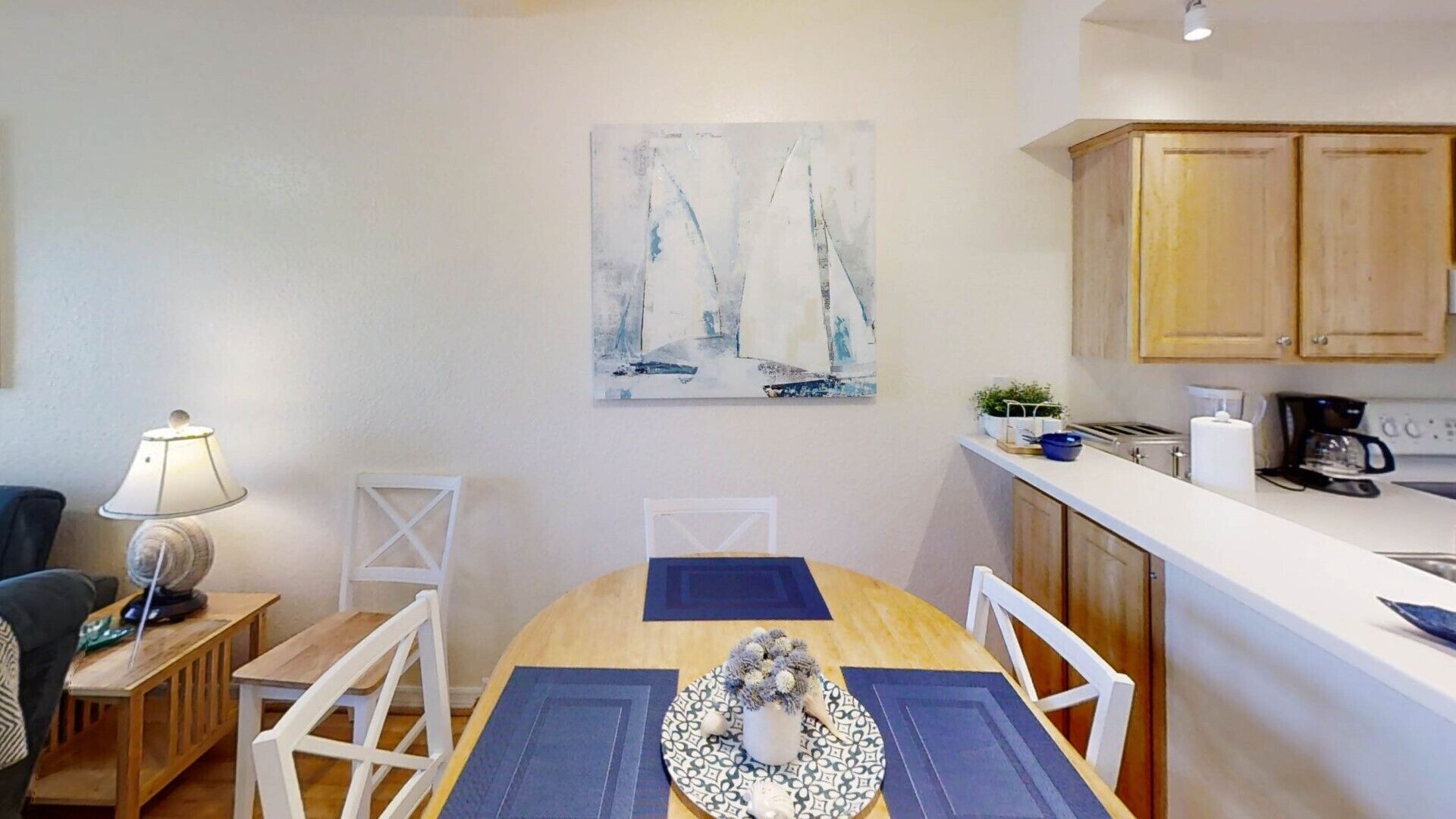 A dining area with a wooden table, white chairs, a blue placemat, and a decorative vase. A painting of sailboats is on the wall.