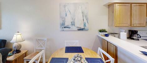 A dining area with a wooden table, white chairs, a blue placemat, and a decorative vase. A painting of sailboats is on the wall.