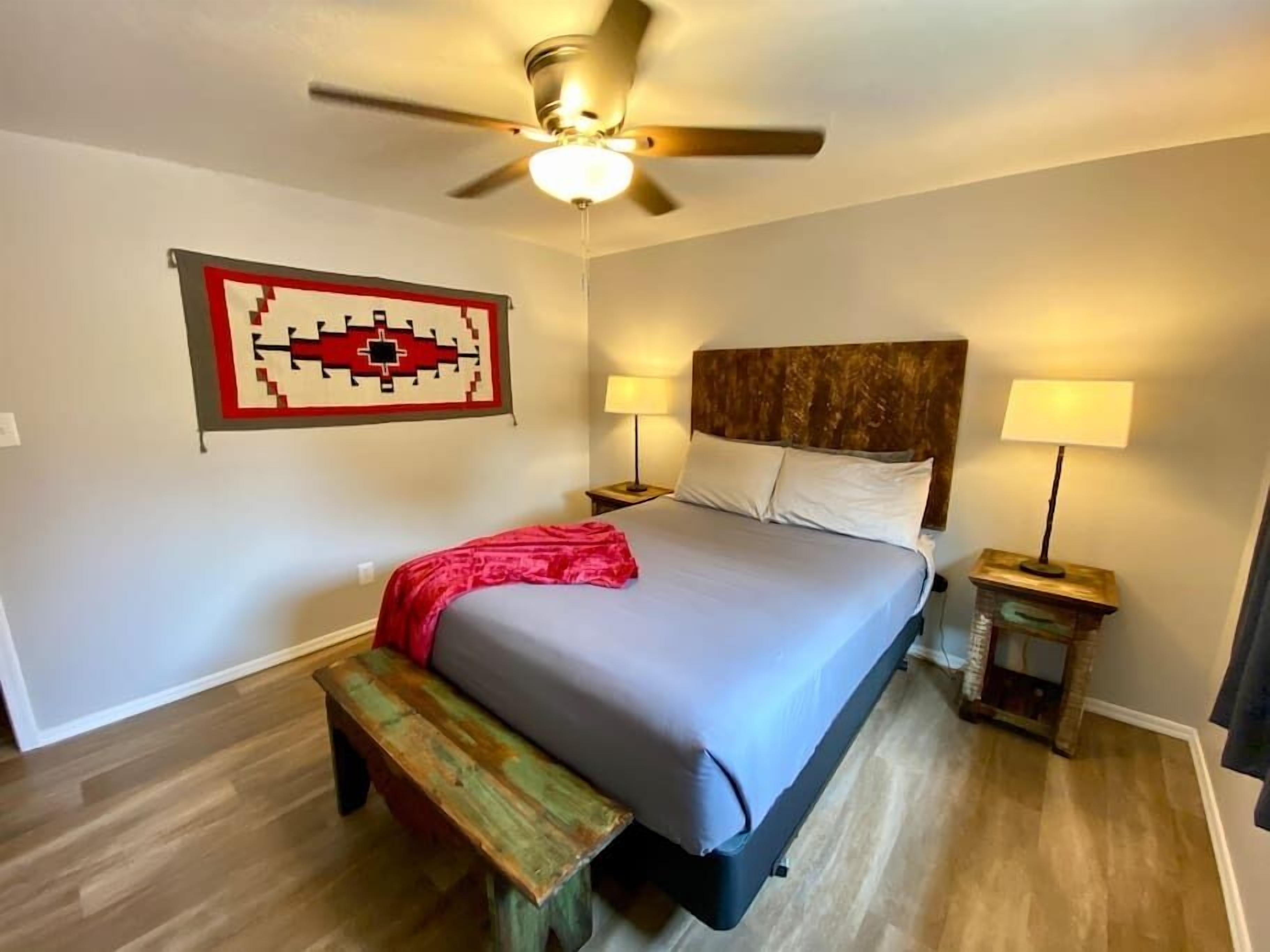 A bedroom with a bed, two bedside tables with lamps, a ceiling fan, a wooden bench, and a wall-mounted artwork.