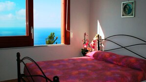 Ein Zimmer mit Bett, einem Meerblick aus dem Fenster, einer Blumenvase und einem gerahmten Bild an der Wand.