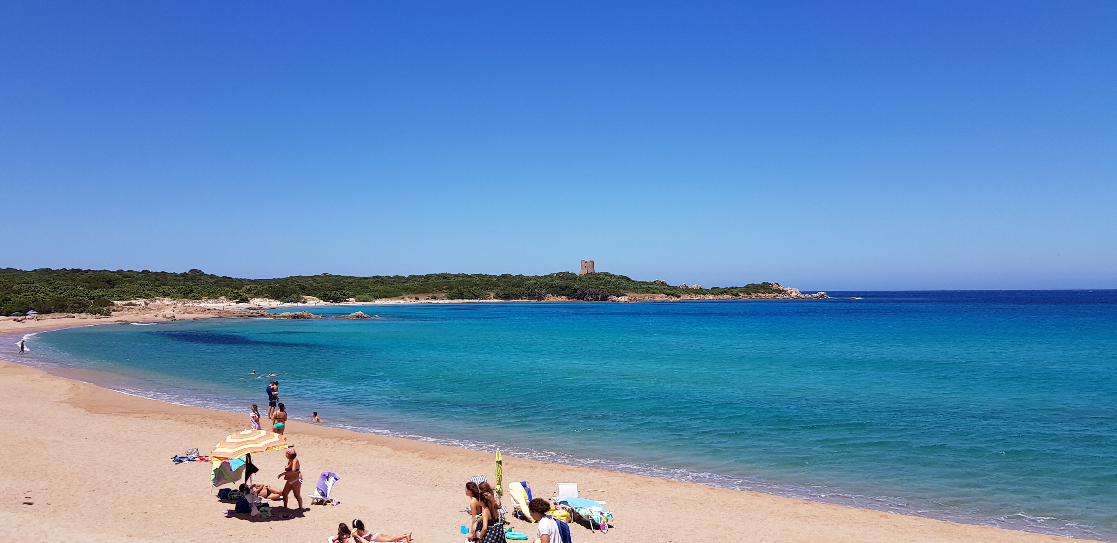 Una playa con agua azul cristalina, orilla de arena y una torre a lo lejos.