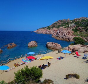 A beach with clear blue water, rocky outcrops, and a few umbrellas.