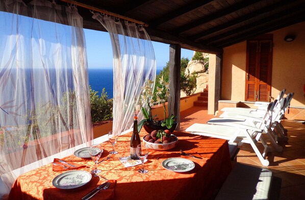 A table set for two with a view of the sea, a bottle of wine, and a bowl of fruit.