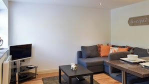 Una sala de estar moderna con un televisor de pantalla plana, una mesa de centro oscura, un sofá gris con cojines coloridos, una pequeña figurita de perro decorativa y un letrero en la pared que dice 'Be our guest'.