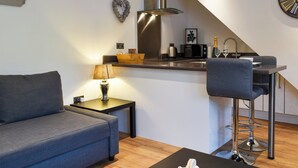 Un espacio habitable compacto con sofá, una mesita con lámpara, kitchenette con microondas y un taburete de bar.