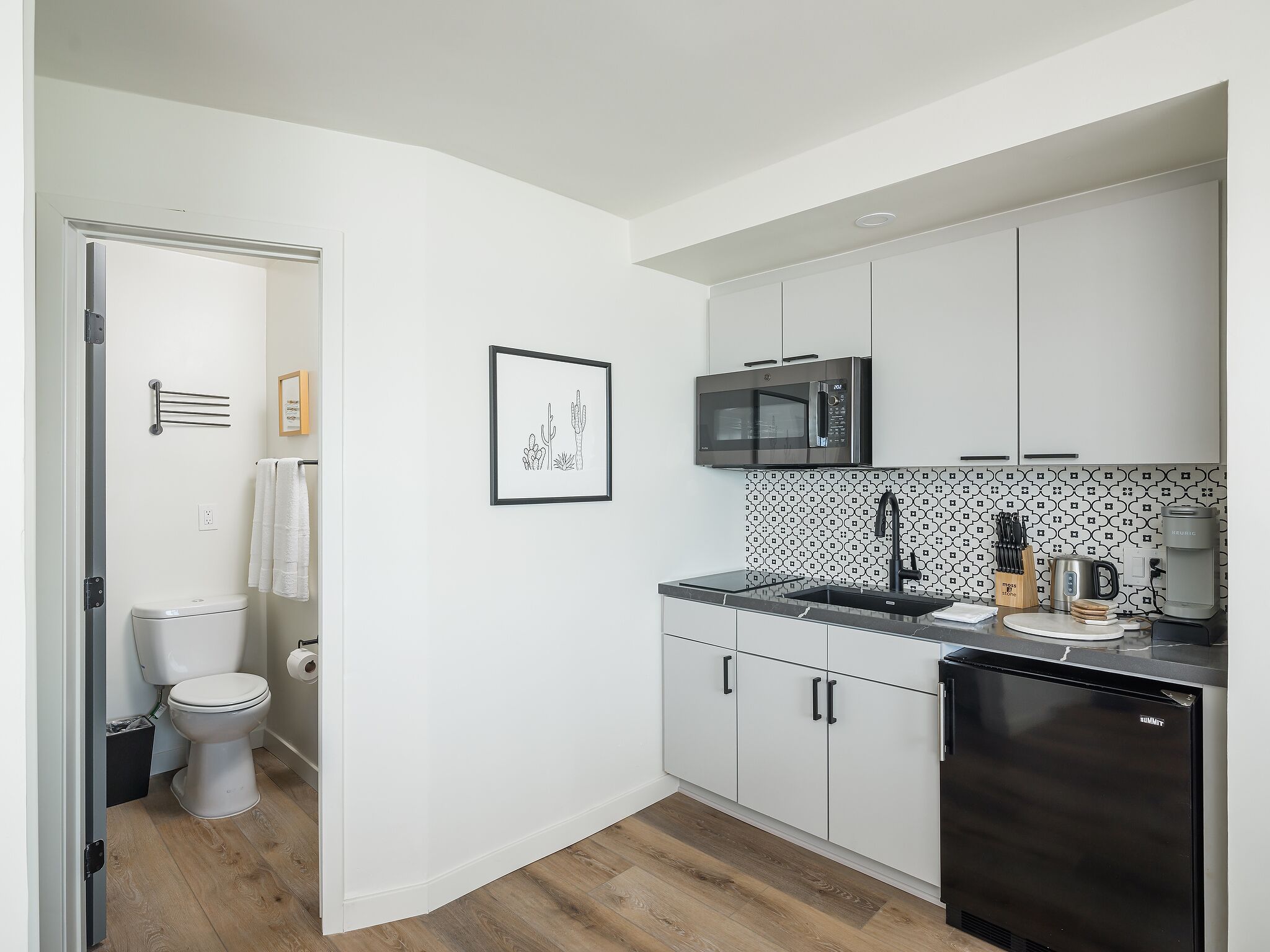 Una cocina compacta con armarios blancos, un refrigerador negro, un microondas y una cafetera.
