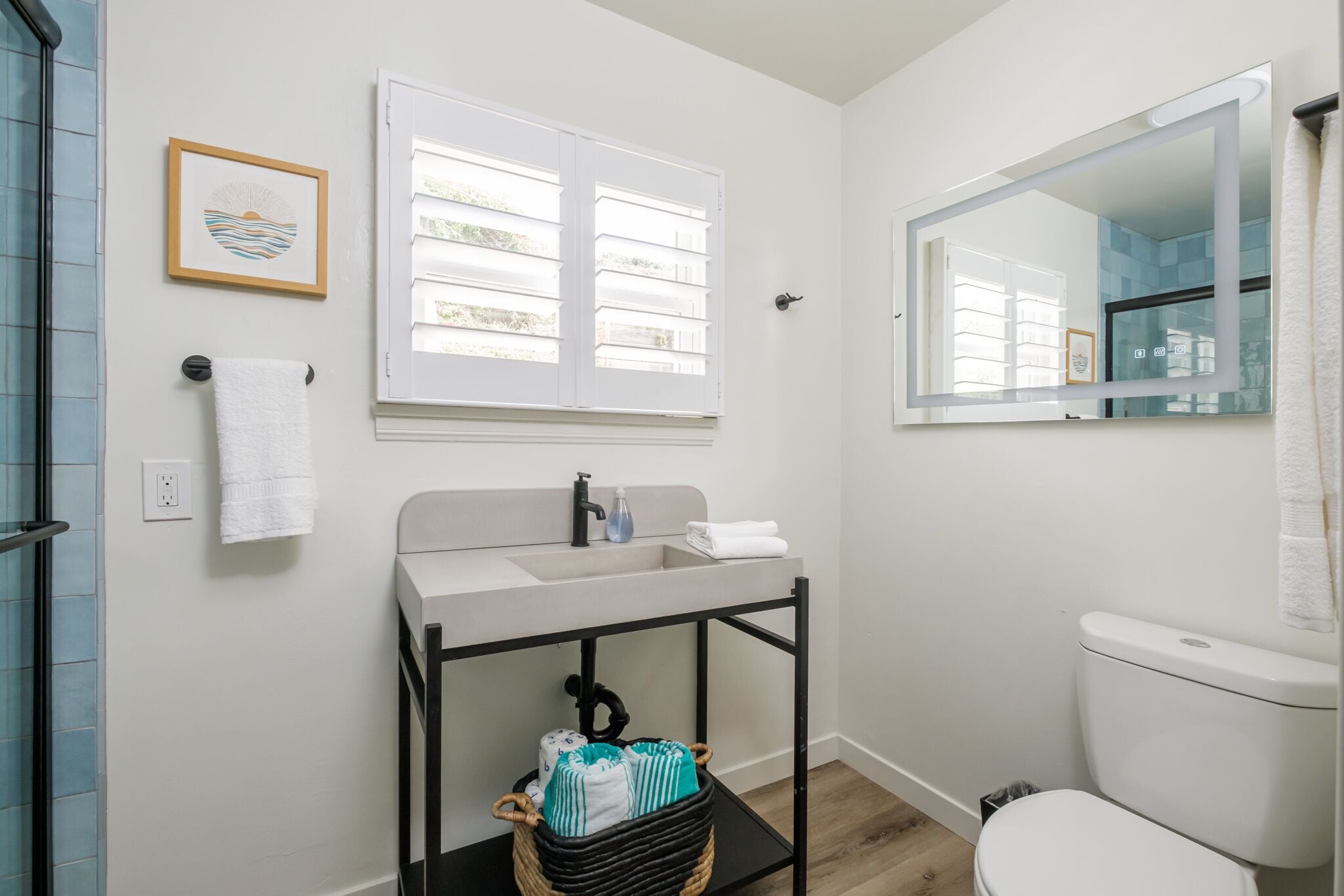 A bathroom with a white sink, a black vanity, a toilet, a window with blinds, and a framed picture on the wall.