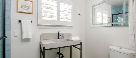 A bathroom with a white sink, a black vanity, a toilet, a window with blinds, and a framed picture on the wall.