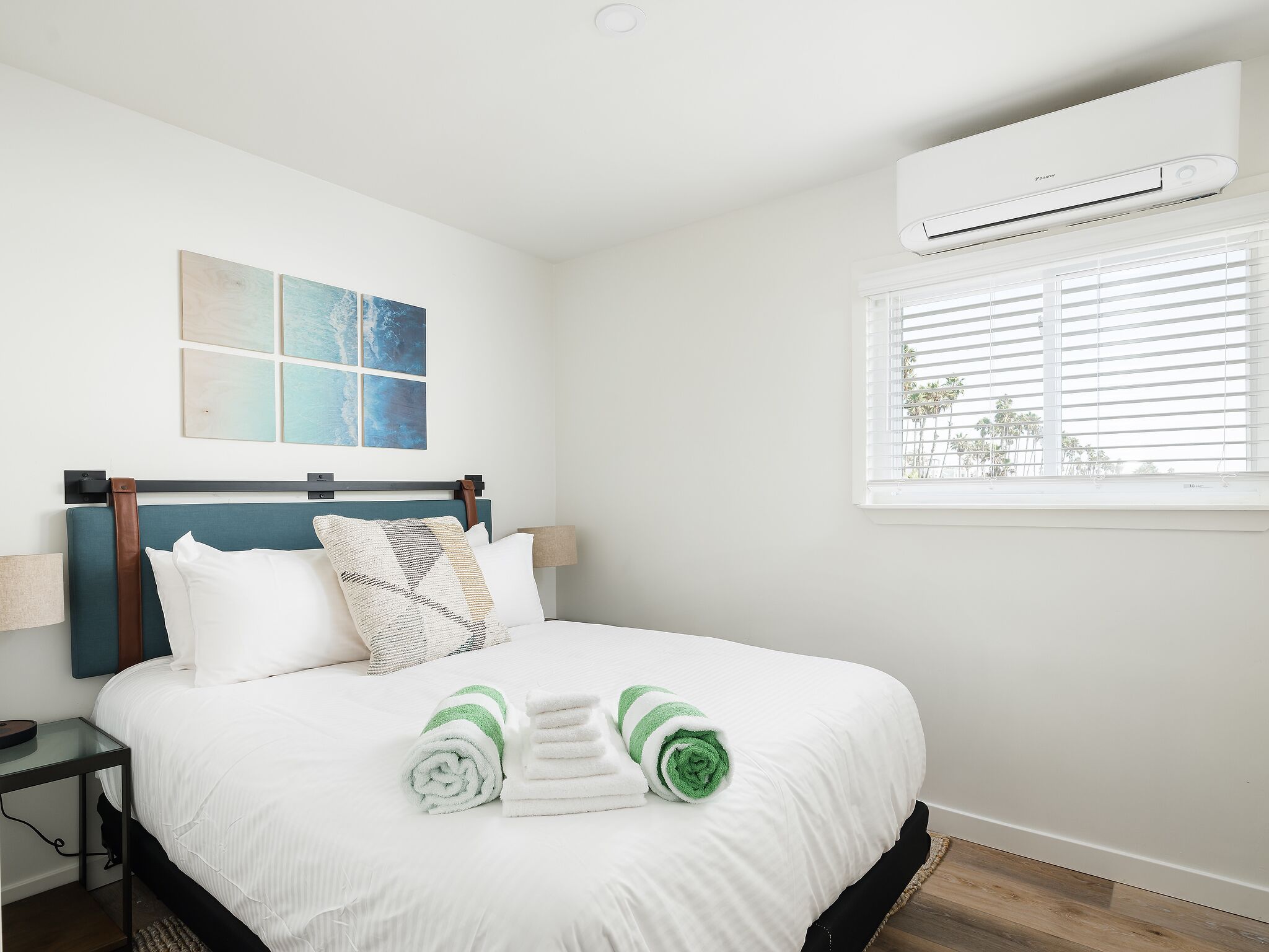 A neatly made bed with white and green towels, a blue headboard, and a window with blinds.