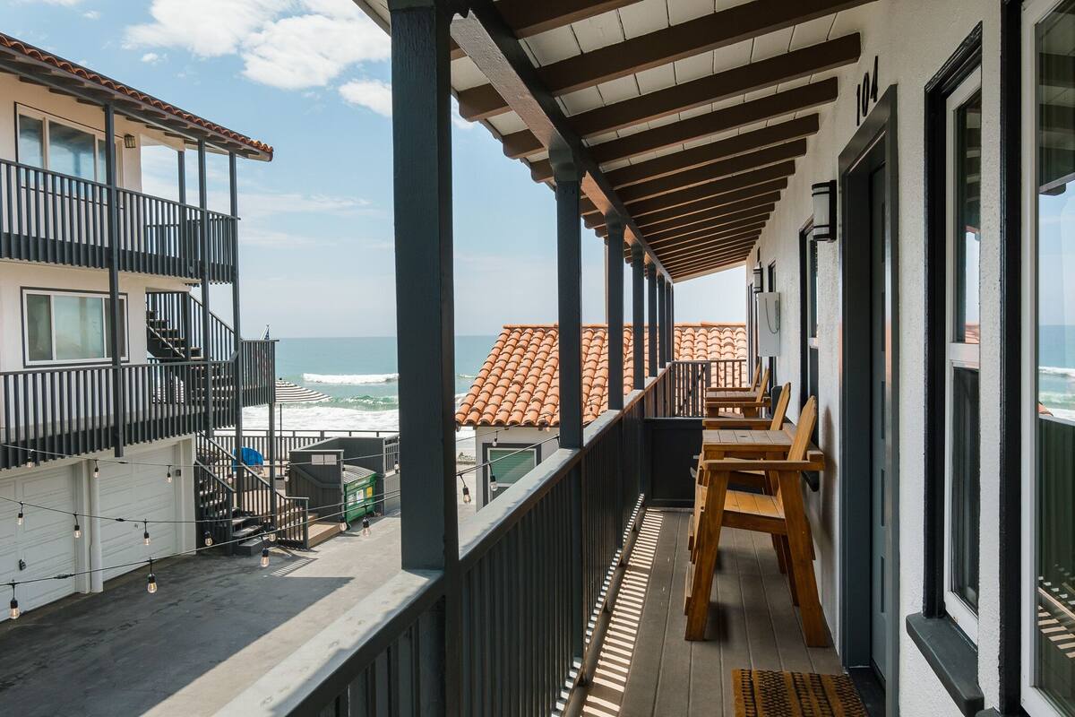 A balcony with wooden furniture, a view of the ocean, and a building with a tiled roof.