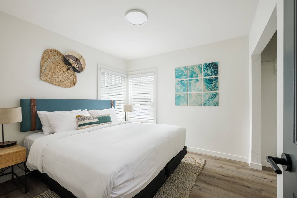 A bedroom with a large bed, a headboard, a bedside table, a lamp, a woven wall hanging, a window with blinds, and a wall with a blue abstract painting.