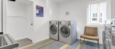 A laundry room with two front-loading washing machines, a chair, and a window.