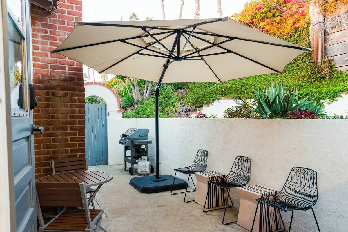 A patio area with a brick wall, a wooden table, two chairs, a grill, and an umbrella.