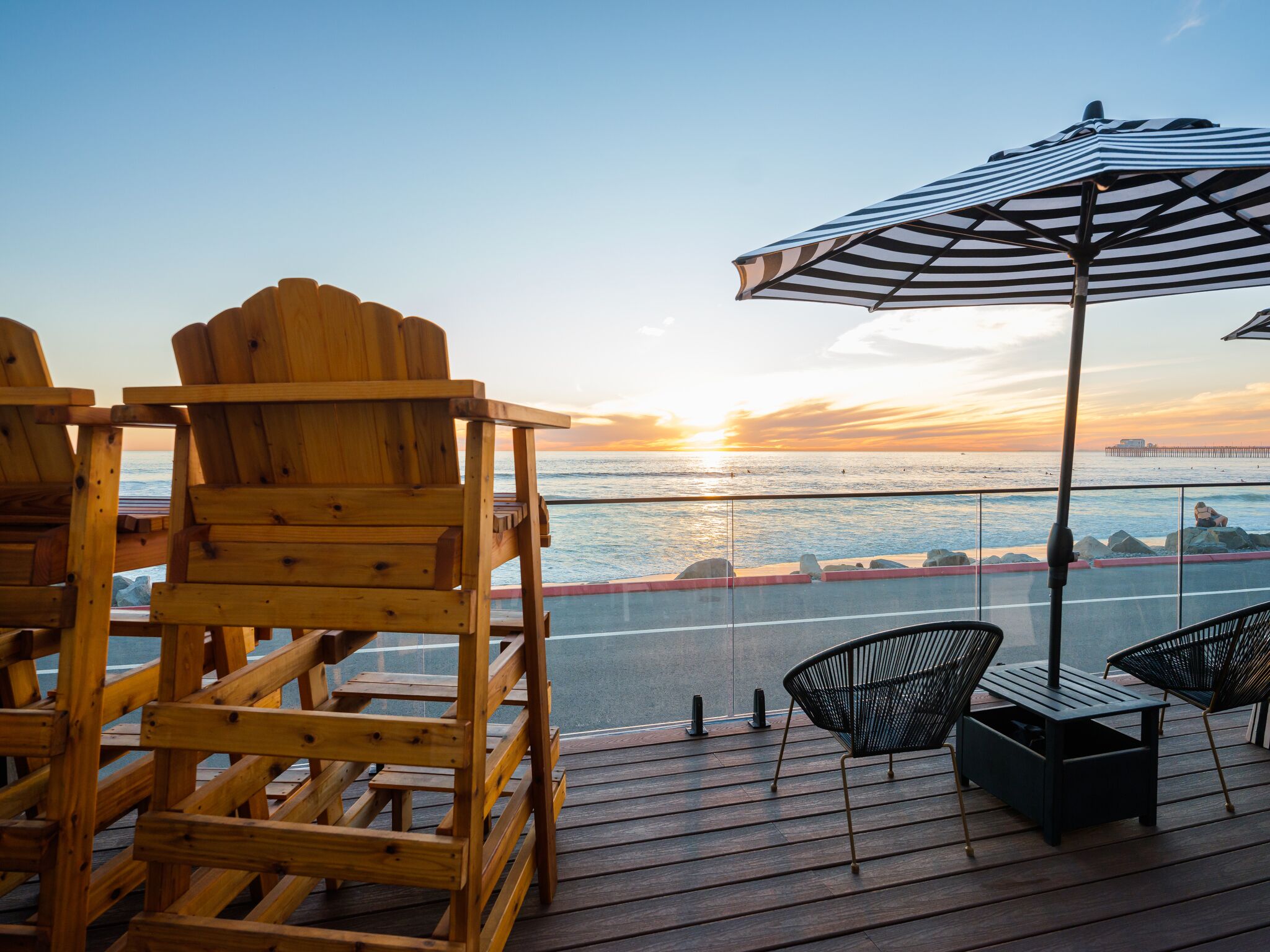 Terraza de madera con sillas Adirondack y una silla de metal negro, con vista a una puesta de sol sobre el agua.