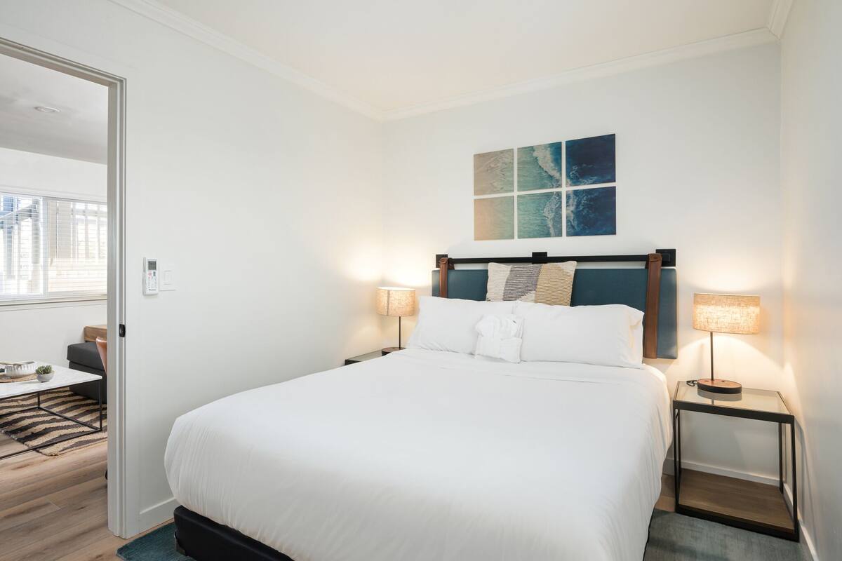 A modern hotel room with a large bed, bedside tables with lamps, a view of the city through a window, and a painting on the wall.