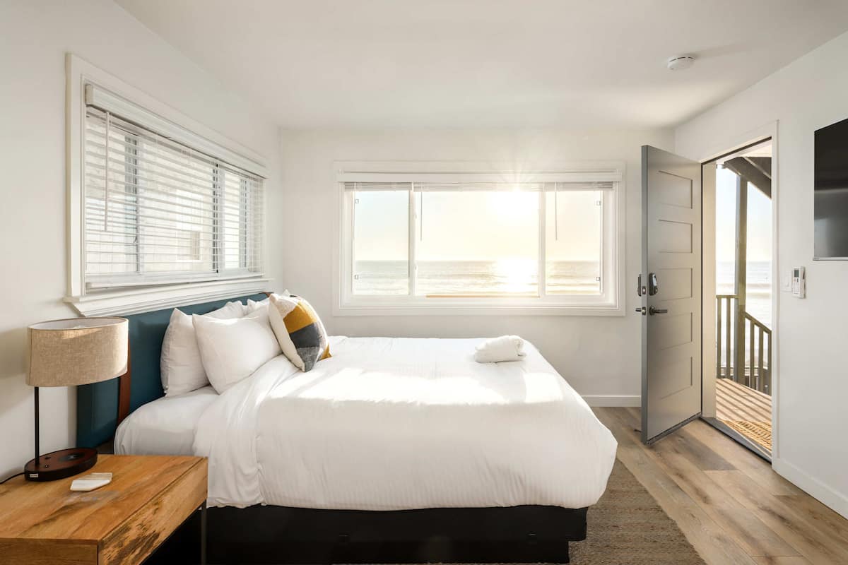 A bedroom with a large bed, a bedside table with a lamp, a window with a view of the beach, and a door leading to a balcony.