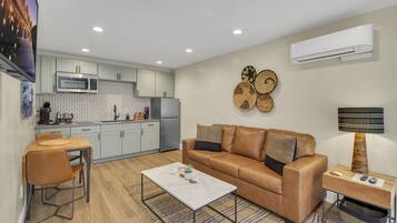 A modern living room with a brown leather sofa, a wooden coffee table, and a kitchen area with light-colored cabinets and a microwave.
