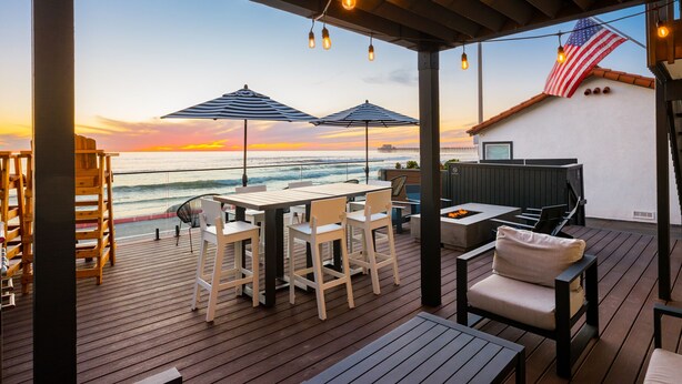 Une terrasse en bois avec coin salon extérieur, un barbecue et une vue sur l’océan au coucher du soleil.