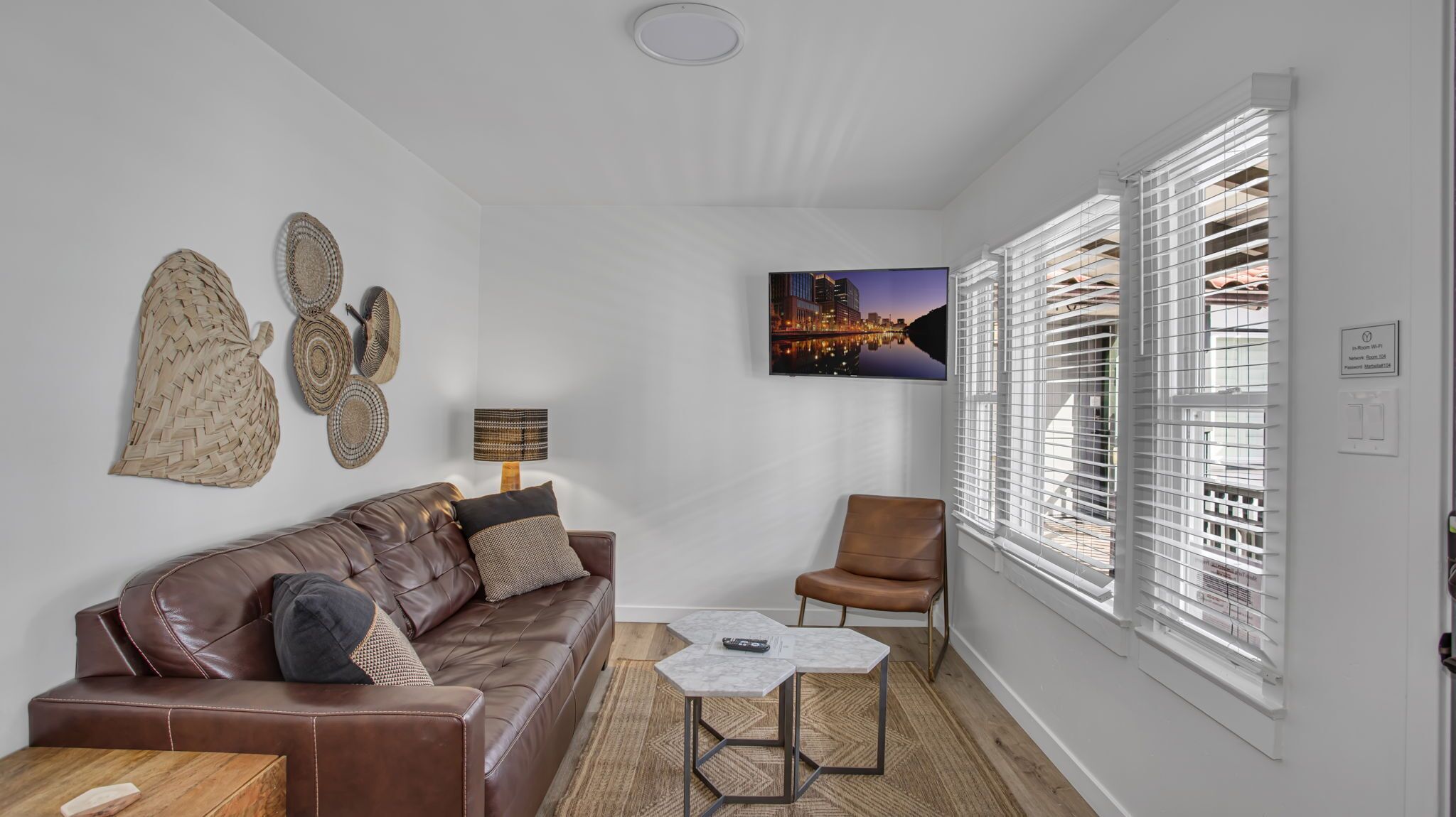 A modern living room with a leather sofa, a coffee table, and a chair. There is a wall-mounted TV and a window with blinds.
