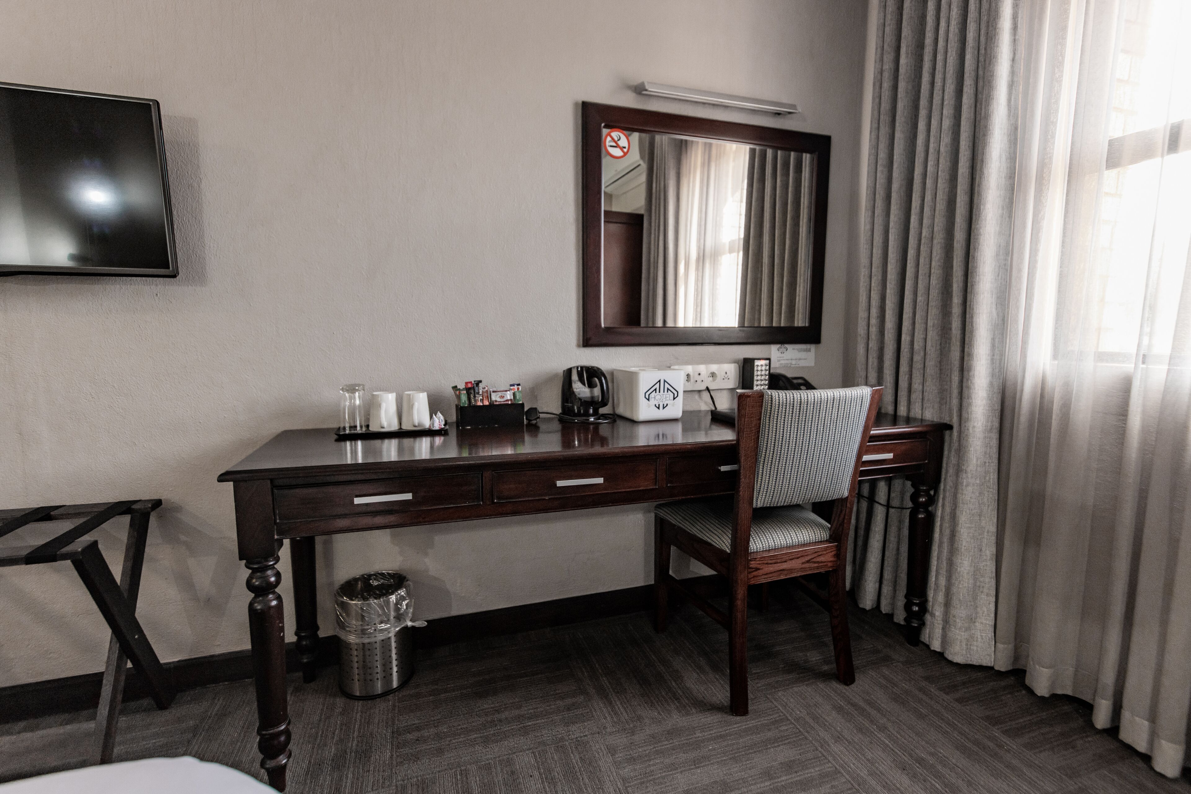 Een hotelkamer met een houten bureau, een stoel, een televisie, een spiegel, een radiator en een raam met gordijnen.