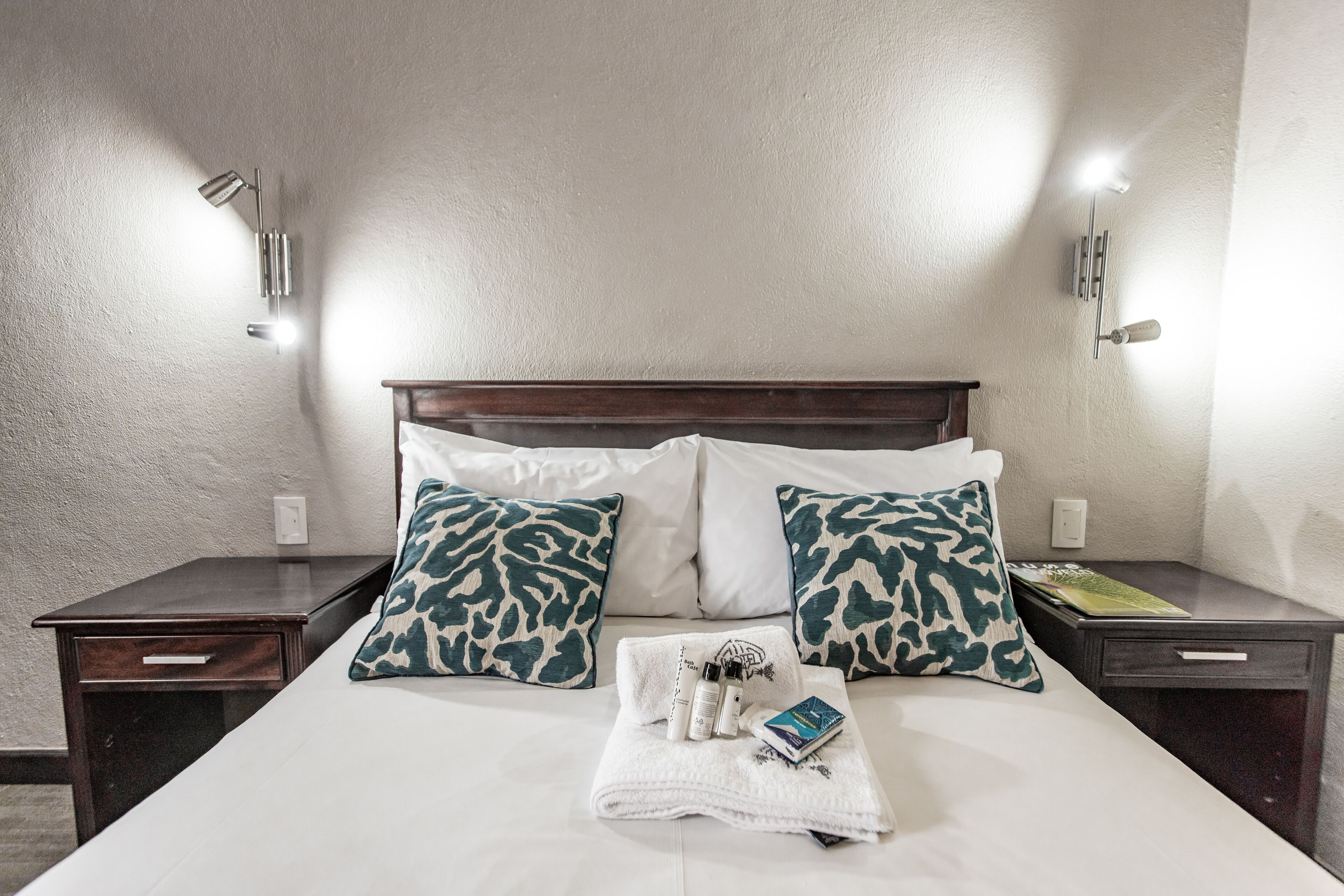 Een net opgemaakt bed met een wit en groen gestreept kussen, een witte handdoek en een boek. Er staan twee nachtkastjes, waarvan er één een boek en een fles op heeft staan.