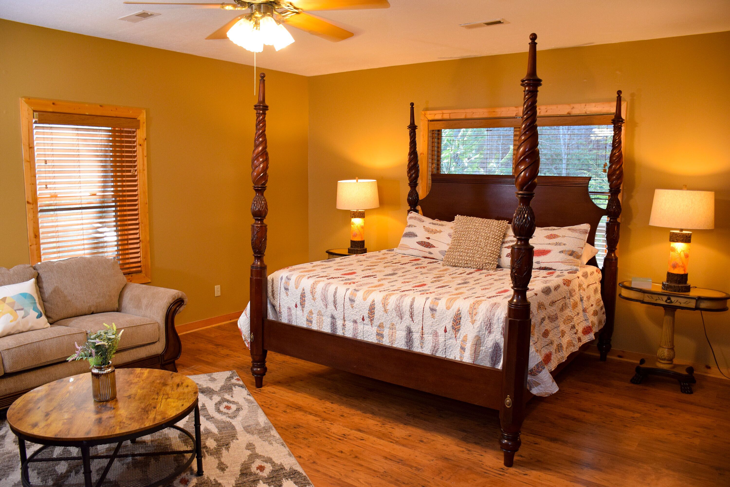 A four-poster bed with a floral bedspread, two bedside tables with lamps, a wooden floor, a sofa with cushions, and a ceiling fan.