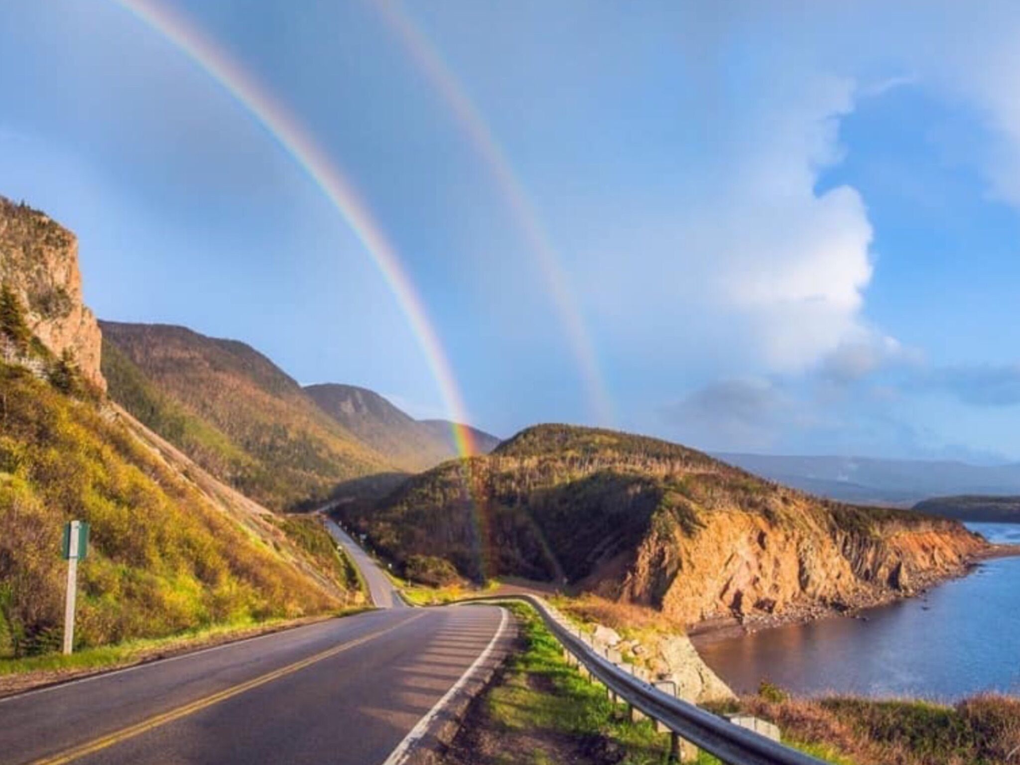 Une route sinueuse traversant un paysage montagneux, avec une étendue d’eau d’un côté et un arc-en-ciel dans le ciel.