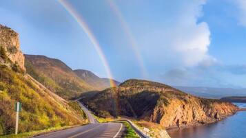 Une route sinueuse traversant un paysage montagneux, avec une étendue d’eau d’un côté et un arc-en-ciel dans le ciel.