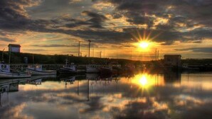 Una marina con barche ormeggiate, acque calme che riflettono il cielo e un tramonto sullo sfondo.