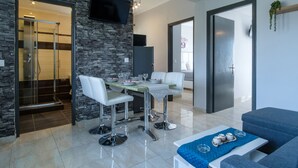 A modern living room with a stone accent wall, a dining area with white chairs, a blue sofa, and a flat-screen TV mounted above the fireplace. - Akis Villa Studios & Apartments with Pool (Corfu)