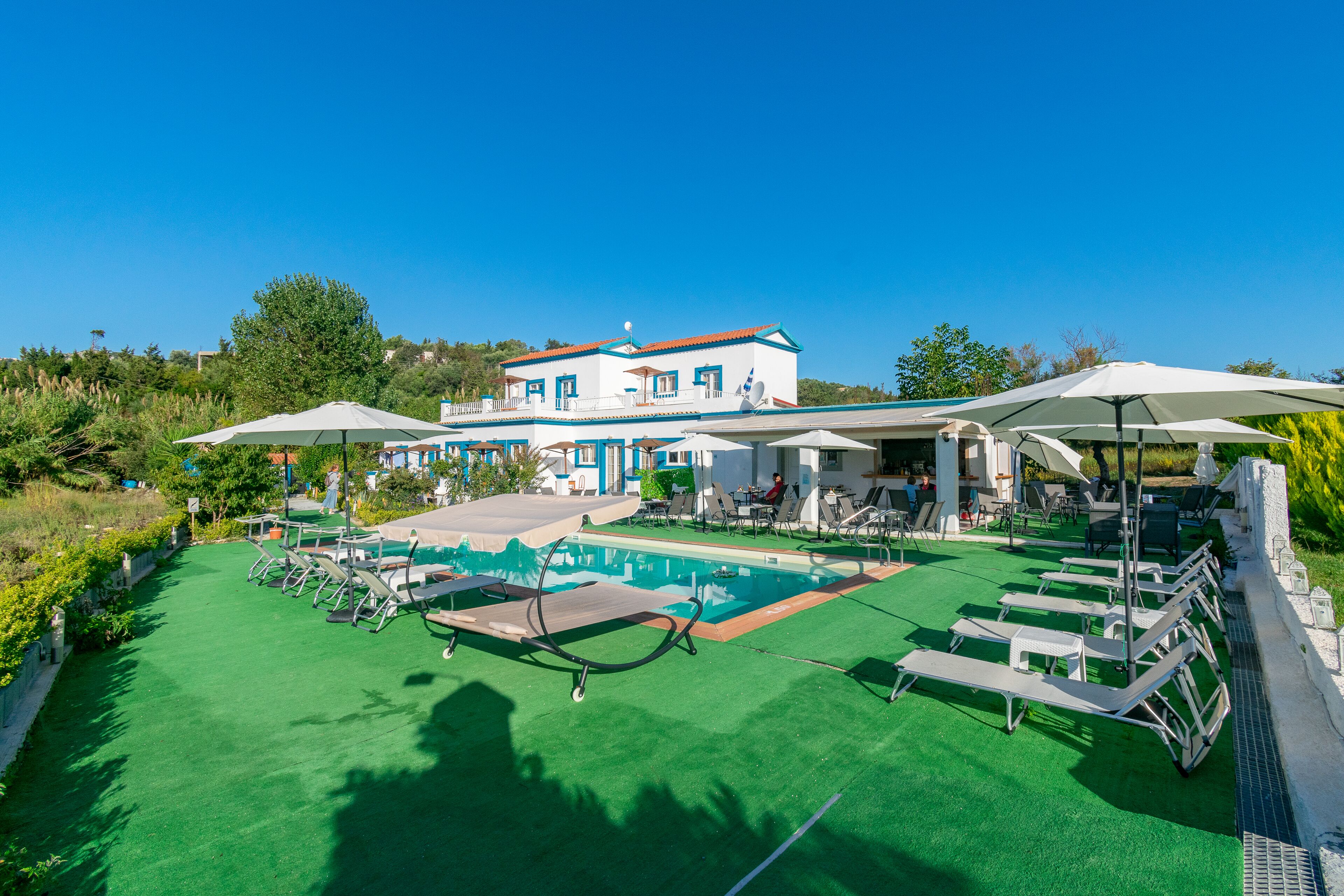 Uma área de piscina com espreguiçadeiras e guarda-sóis, um edifício com fachada azul e branca, e um céu azul e límpido.