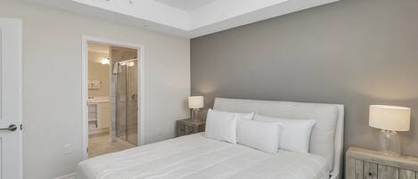 Un dormitorio con una cama grande, dos mesitas de noche con lámparas, una puerta que da al baño y una pared de acento gris.
