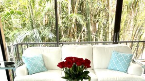 A white sofa with blue cushions, a glass coffee table with decorative items, and a view of lush greenery outside. - Beach Condo located in the Heart of Olde Naples (Naples)