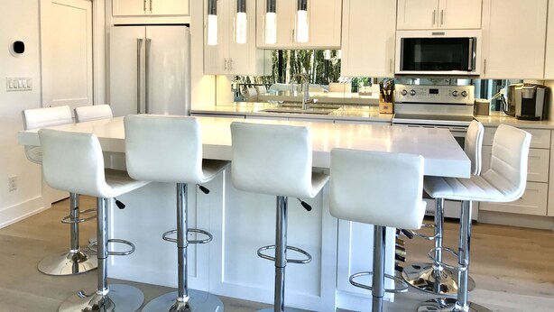 A modern kitchen with white cabinetry, a central island with bar stools, stainless steel appliances, and pendant lighting. - Beach Condo located in the Heart of Olde Naples (Naples)