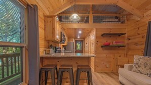 A wooden cabin interior with a kitchen, bar stools, a sofa, and a window with blinds. - Peggers Luxury Tiny Cabin at Lookout - Loaded (Chattanooga)