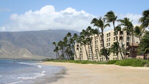 Un hôtel situé en bord de mer, doté d’une plage de sable, de palmiers et offrant une vue sur les montagnes.