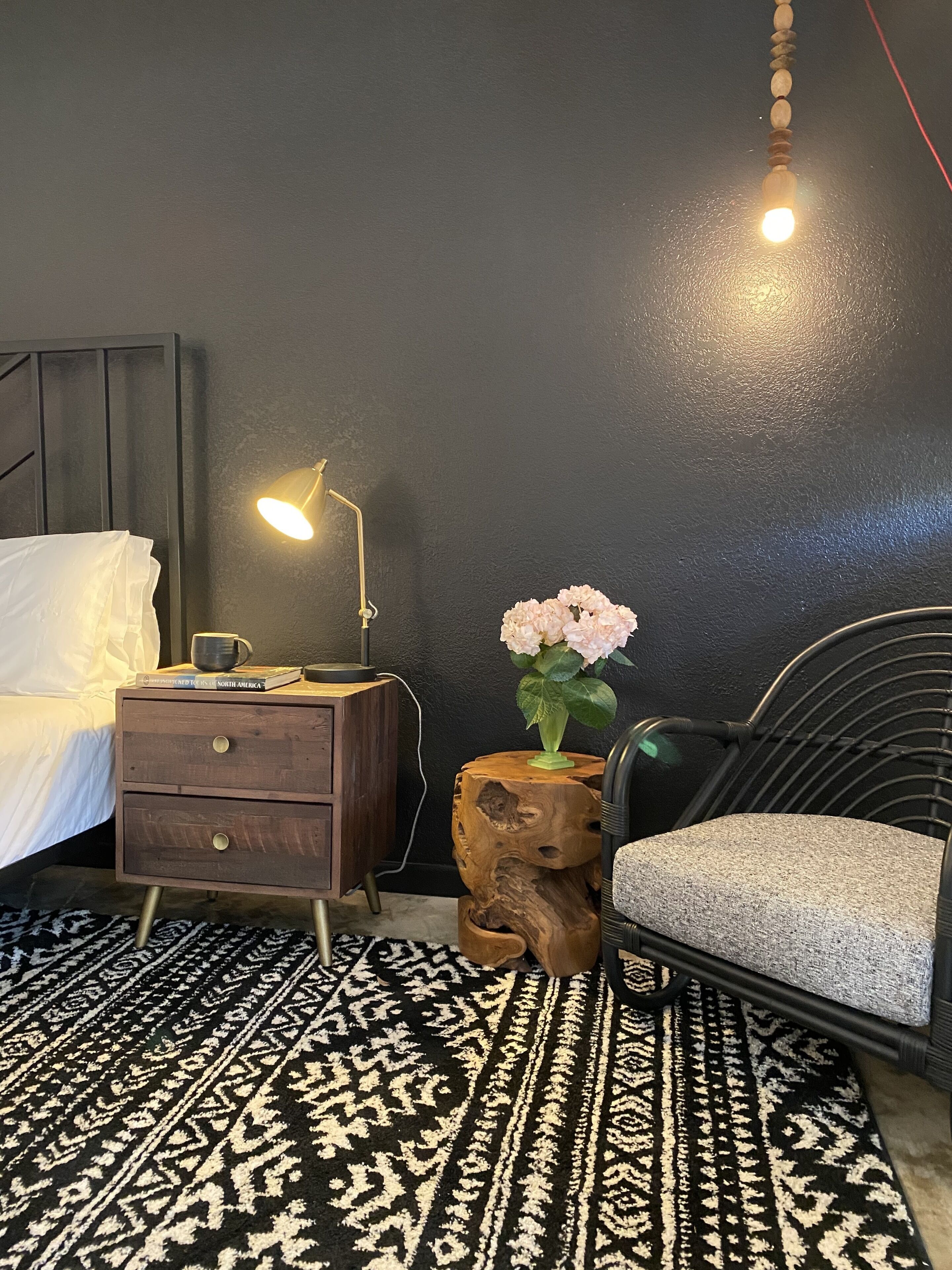 A bedroom with a bed, bedside table with a lamp, a chair, a wooden stool with a vase of flowers, and a patterned rug.