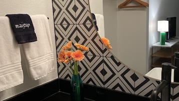 Un baño con lavamanos blanco, un espejo, un perchero con una toalla negra y un jarrón con flores anaranjadas.
