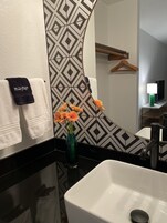 Un baño con lavamanos blanco, un espejo, un perchero con una toalla negra y un jarrón con flores anaranjadas.