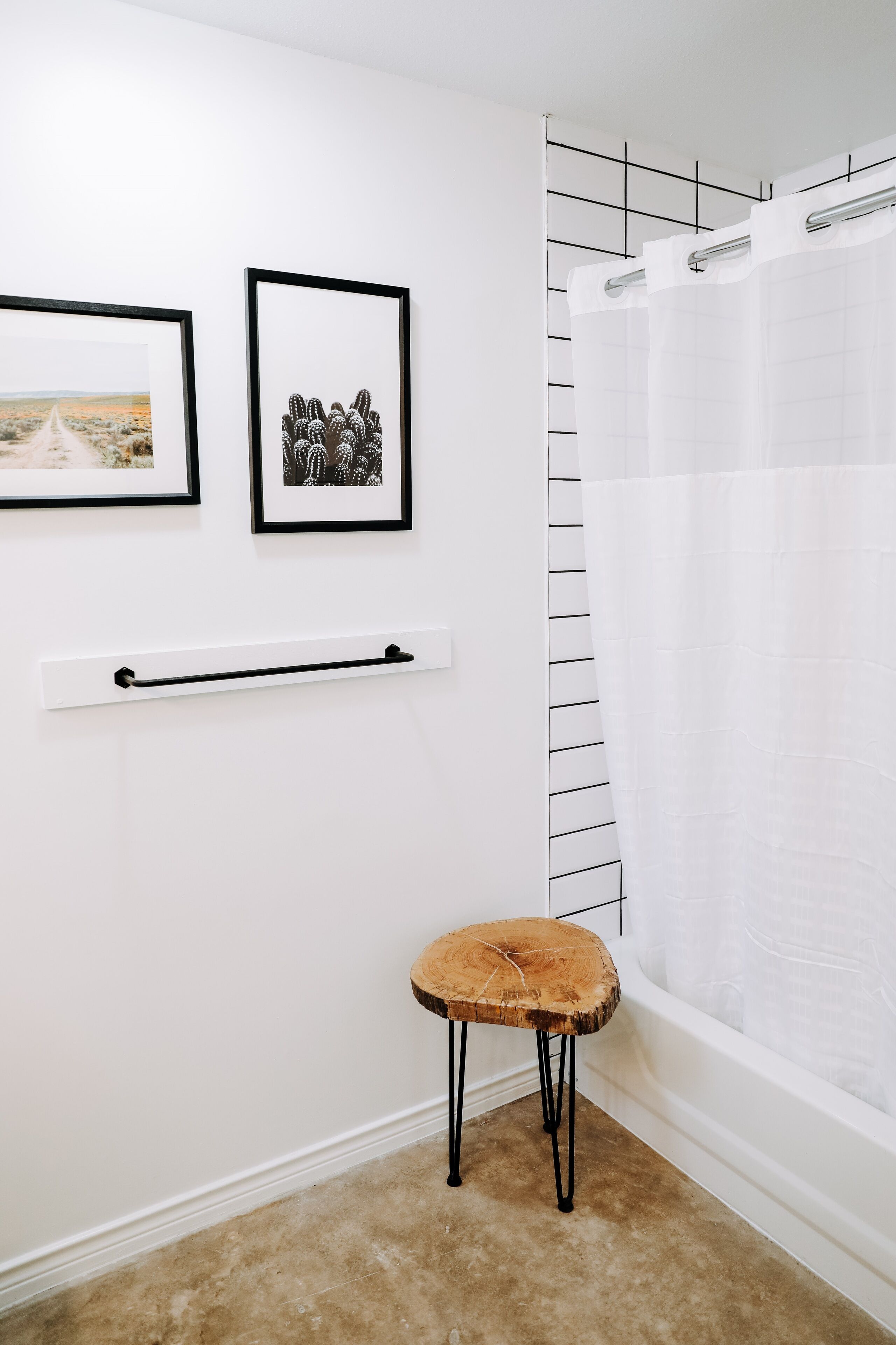 Une salle de bain avec une baignoire blanche, un rideau de douche, un tabouret en bois et deux tableaux encadrés accrochés au mur.