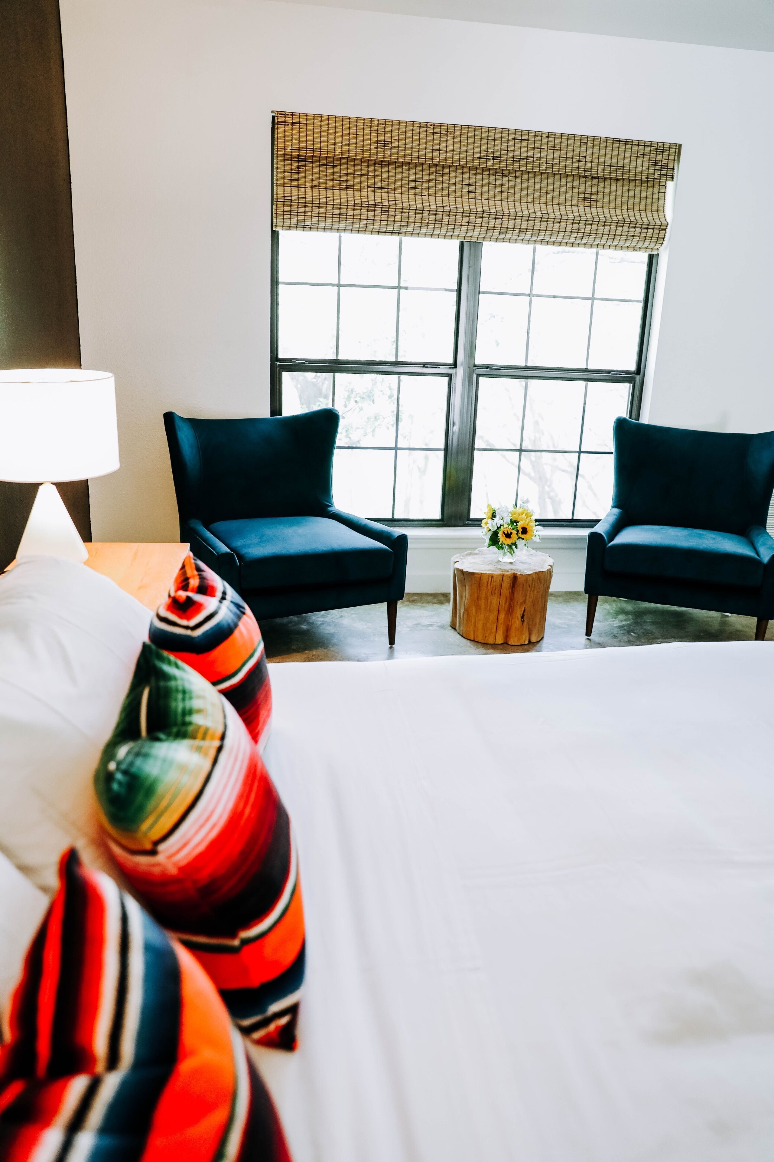 Ein Hotelzimmer mit einem großen Bett, zwei dunklen Sesseln, einem runden Holztisch mit Blumenvase und einem Fenster mit Gittermuster.
