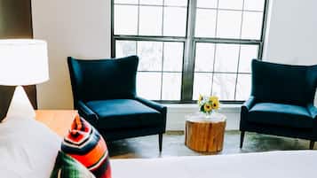 Ein Hotelzimmer mit einem großen Bett, zwei dunklen Sesseln, einem runden Holztisch mit Blumenvase und einem Fenster mit Gittermuster.