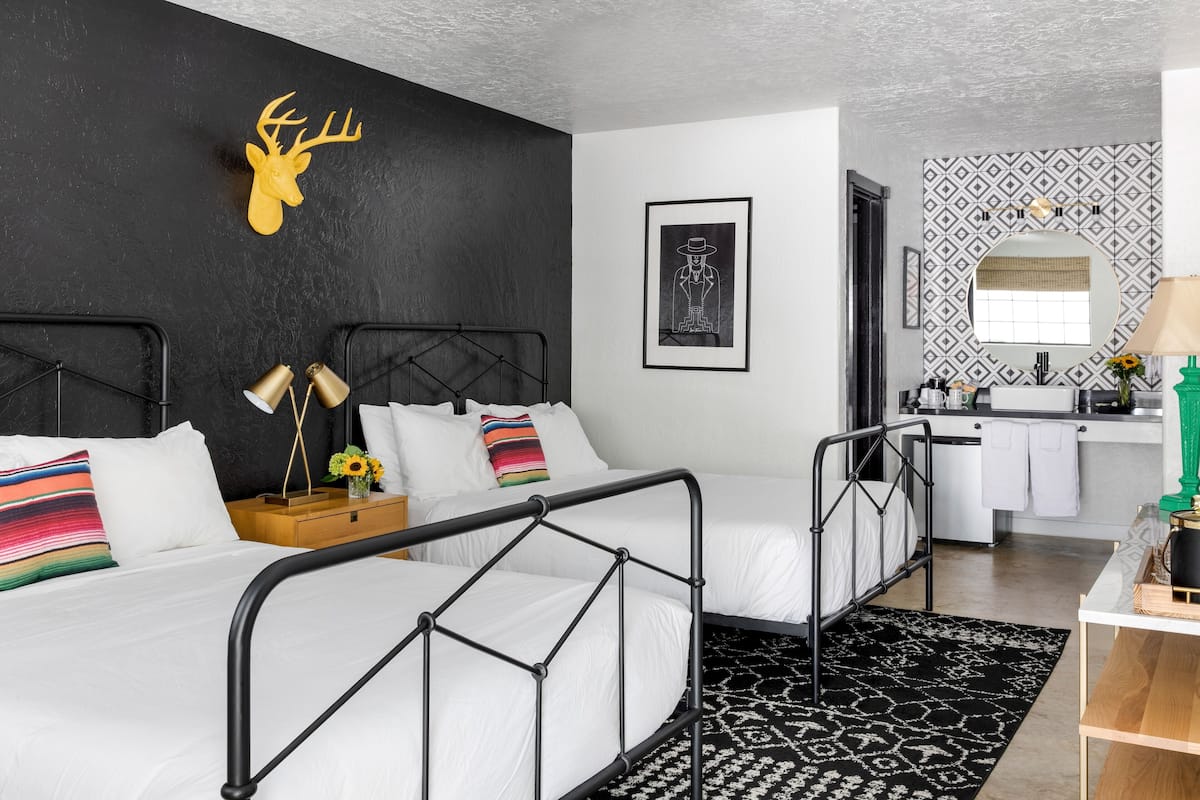 Quarto moderno com esquema de cores preto e branco, apresentando uma cama com estrutura de metal, um chifre de veado montado, uma moldura com imagem e uma área de cozinha com pia e bancada visíveis.