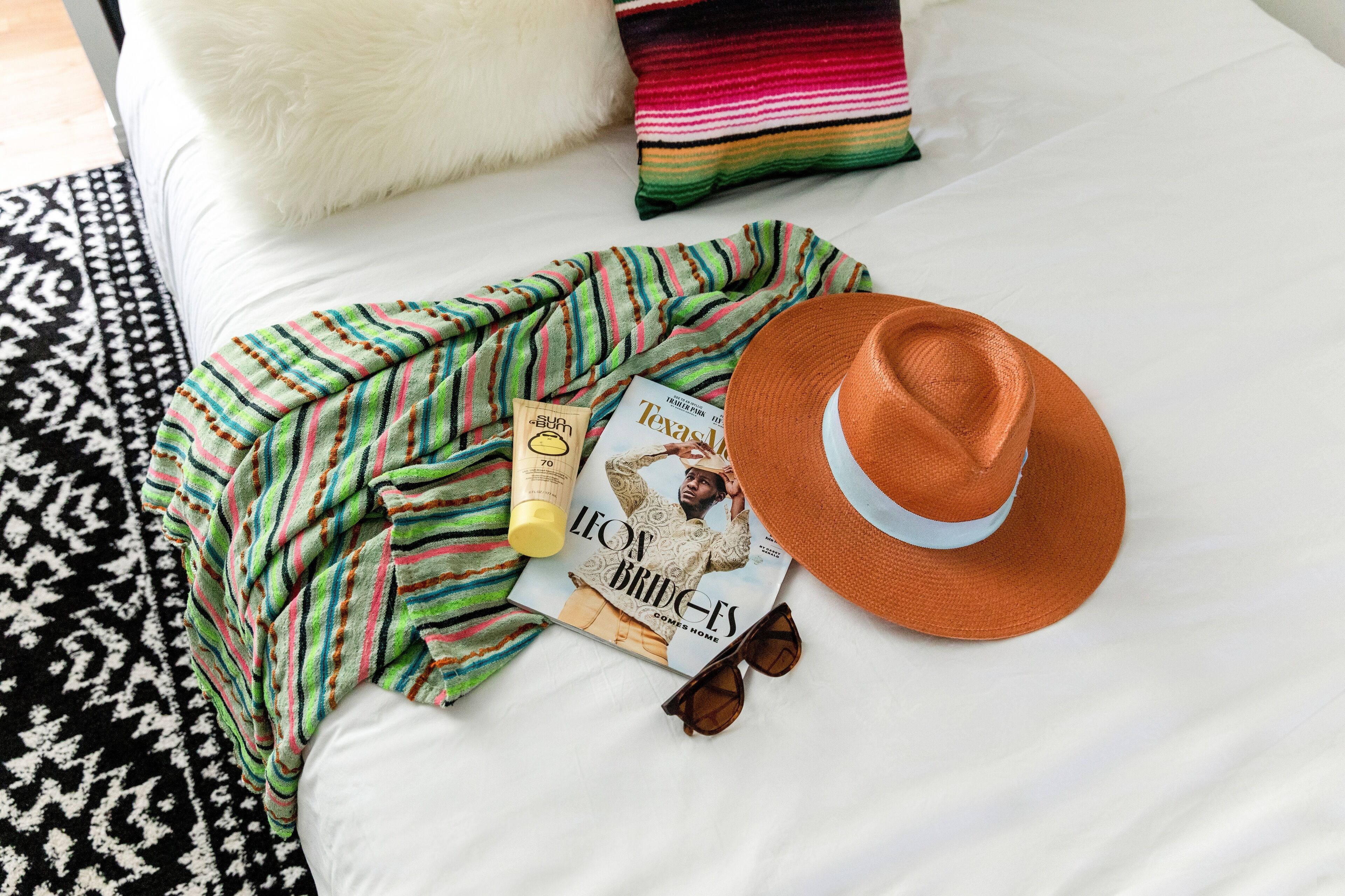 A bed with a white sheet, a striped blanket, a magazine, a bottle, and a hat.
