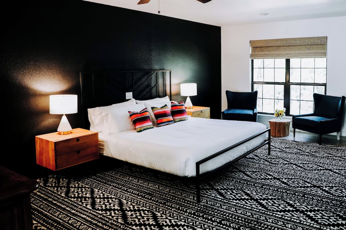Quarto de hotel moderno com uma cama grande, duas mesas de cabeceira com luminárias, um ventilador de teto, uma janela com vista e um tapete estampado.