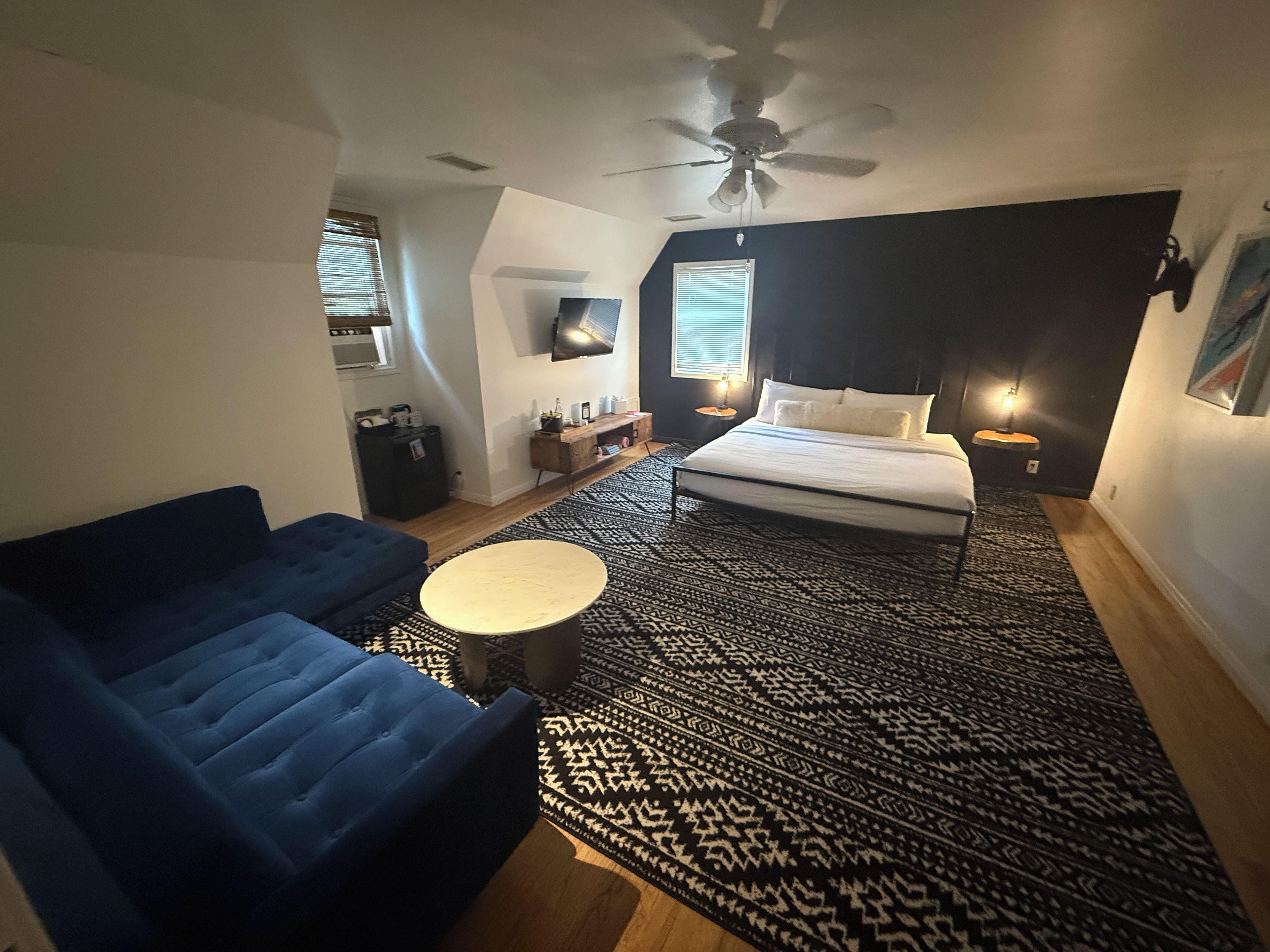 A bedroom with a bed, a blue sofa, a patterned rug, a ceiling fan, and a TV on the wall.