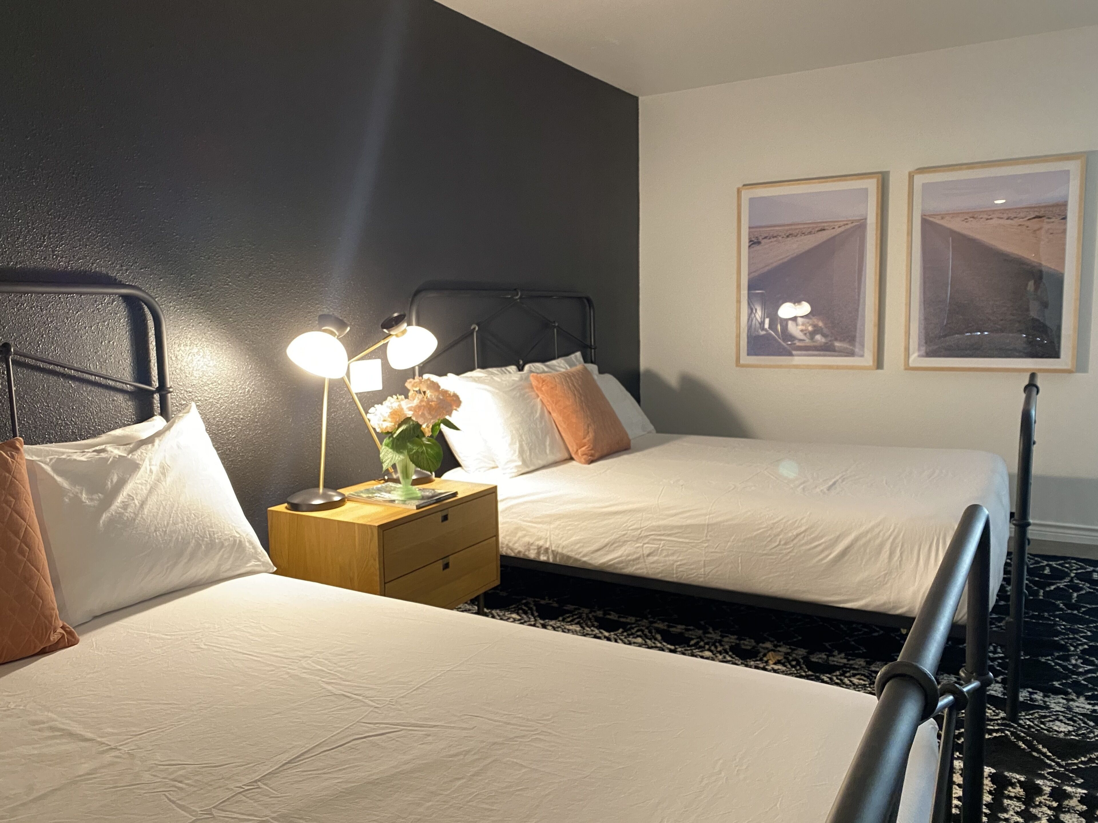 A hotel room with two beds, a nightstand with a lamp, and two framed pictures on the wall.