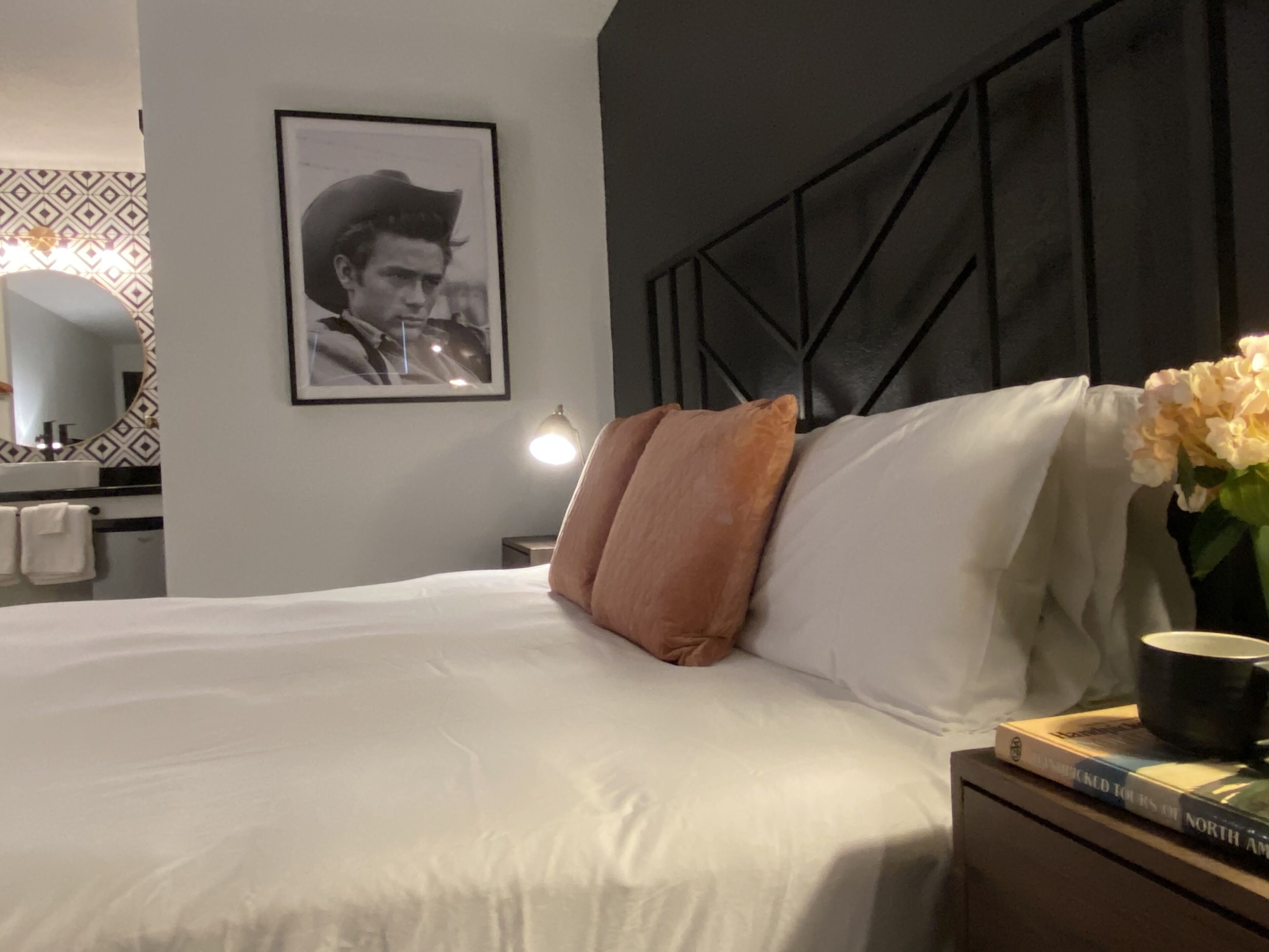 Une chambre d’hôtel avec un lit, une table de chevet, un tableau encadré au mur, un vase avec des fleurs et un livre sur la table de chevet.